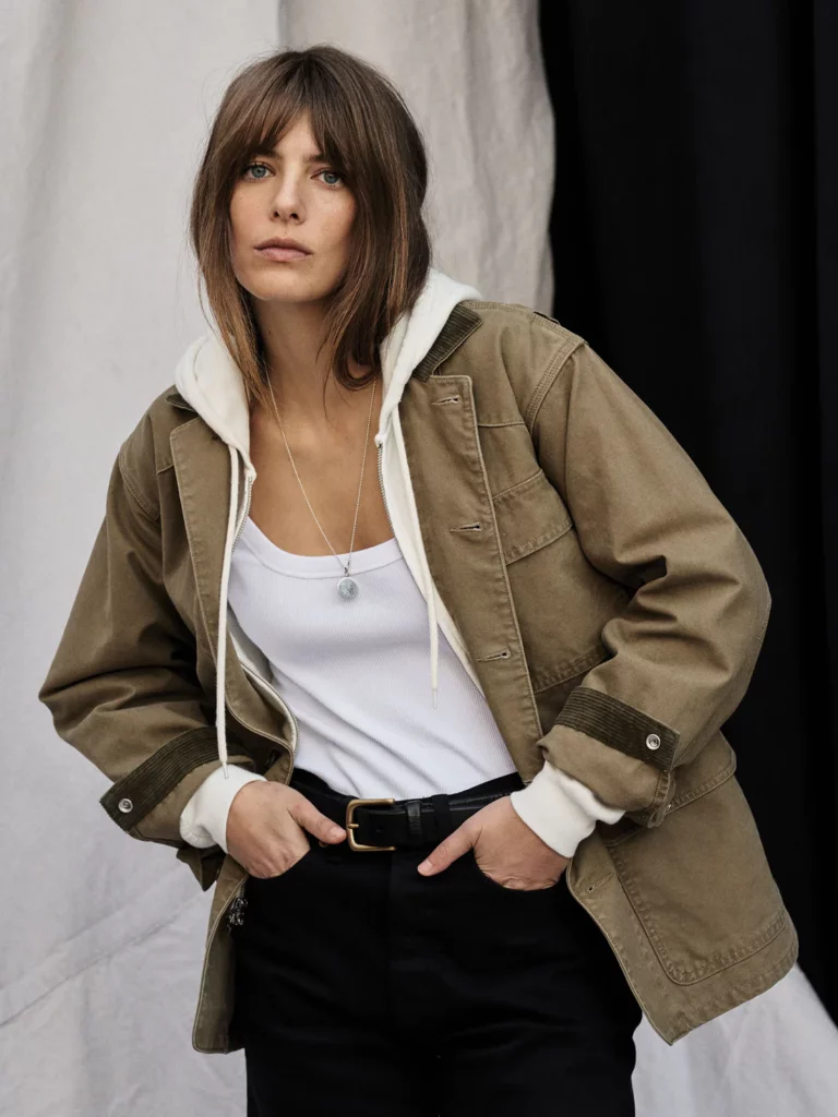 A brown-haired woman in a tan jacket with a hood and white t shirt. 