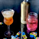 A stemmed glass with a light cola colored cocktail, a gold cocktail shaker and a mason jar with a bubble gum pink vodka, surrounded by bubble gum and bubble gum wrappers