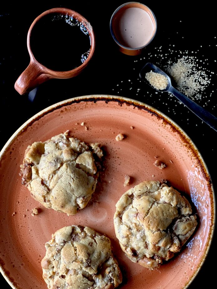Three large Snickers Levain Bakery cookies on a plate with a cup of coffee, a small vessel of creamer, and raw sugar on a spoon