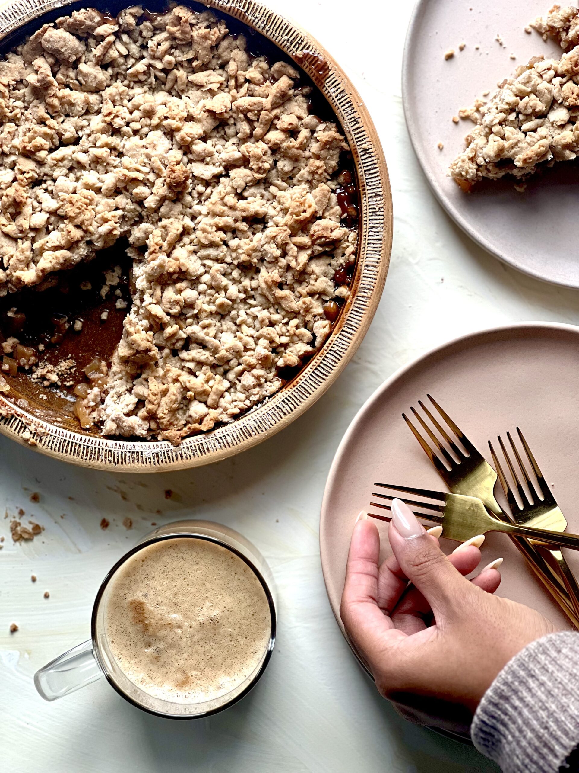 An apple crumble in a round baking dish with a cup of hot cocoa in the left corner, a hand reaching towards a plate of 3 gold forks in the right, and the corner of a piece of the crumble on a plate in the upper right corner.