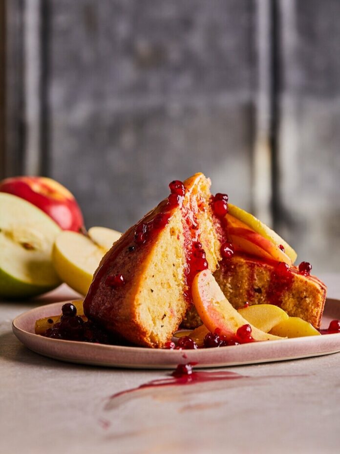 A spongey yellow polenta cake sits on a white plate topped with red spiceberry sauce and fresh apples.