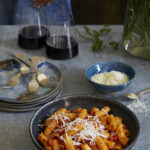 Stanley Tucci Inspired Pasta alla Norma sits in a bowl to the right with two glasses of red wine off to the back left.