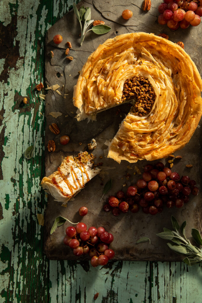 Flat-lay image of a savory cheesecake, roasted grapes on a weathered table.