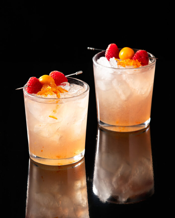 Two cocktails very light orange in color, sit in two rocks glasses. garnished with golden gooseberries and raspberries. The Wiz Cocktail