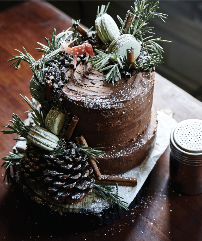 A Whole Grain Apple Cider Layer Cake covered in chocolate frosting and white decor like pinecone and cinnamon sticks.