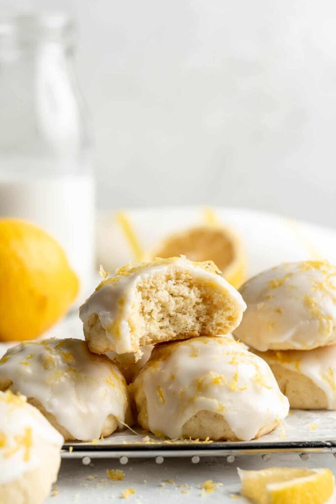 A stack of lemon drop cookies coated in a yellow glaze with a half eaten one on top and a lemon in the background.