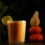 a cocktail orange in color sits in a rocks glass. two pumpkins and a squash are in the background. the cocktail is a pumpkin spice cocktail, a mai tai