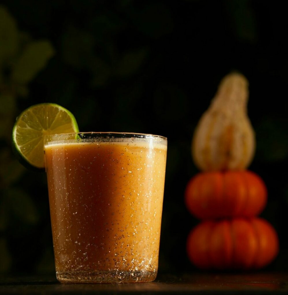 a cocktail orange in color sits in a rocks glass. two pumpkins and a squash are in the background. the cocktail is a pumpkin spice cocktail, a mai tai