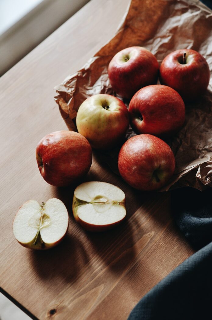 Six apples, with a seventh cut in half, lay on a strip of brown paper on top of a wooden table.