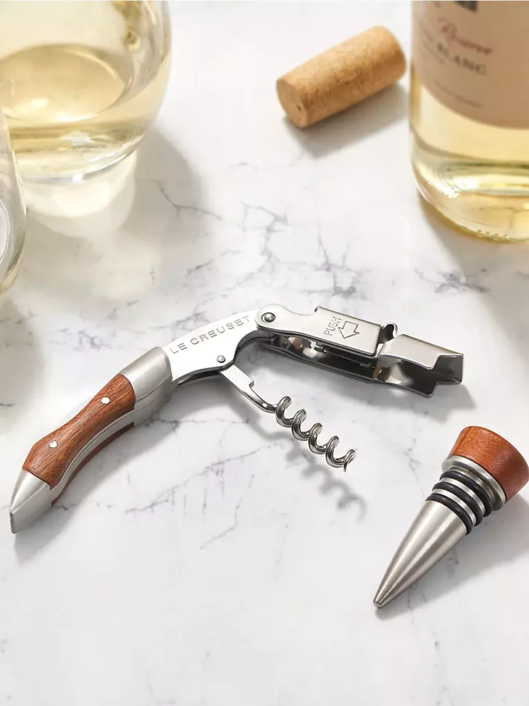 A wine tool and stopper for a home bar lay on a marble table next to a wine cork.