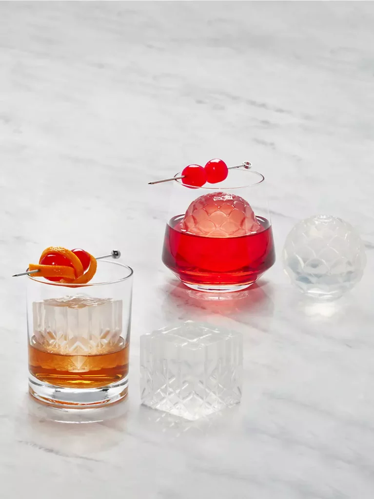 Two cocktail glasses sit on a marble table with fancy square and circle-cut ice cubes in the drink and beside them for a home bar.