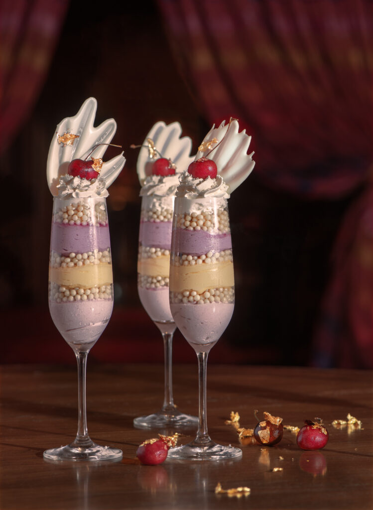 Three identicle champagne flutes filled with mousse and crunchy layers in hues of light purple and vanilla with whipped topping, cherry and a fancy fan garnish on top.