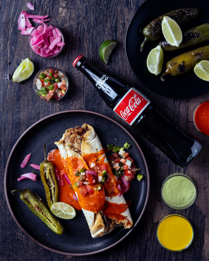 A burrito coated in red sauce sits on a dark plate accompanied by roasted peppers and flower petals. A bottle of coke sits slanted above the plate and another plate can be seen in the top right corner filled with limes.