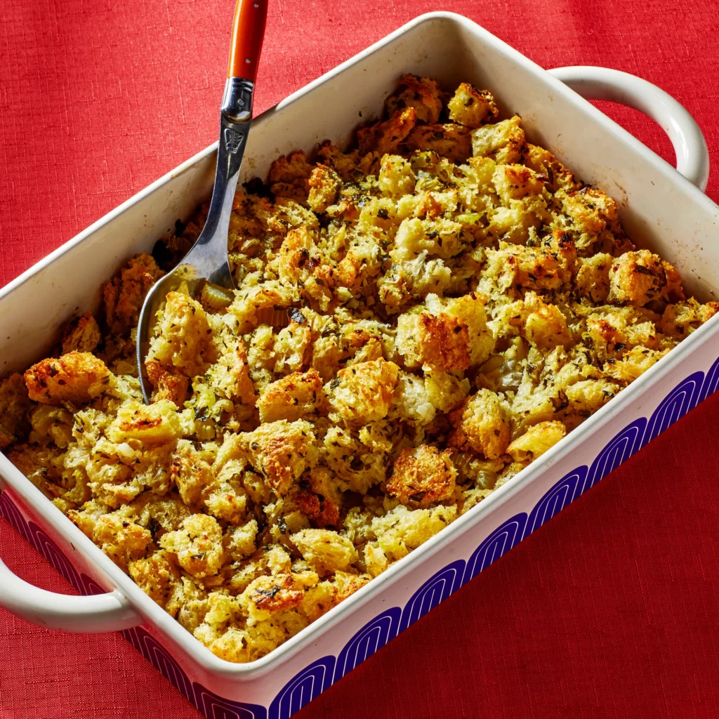 A casserole dish filled with stuffing on a red backdrop with a spoon in the dish.