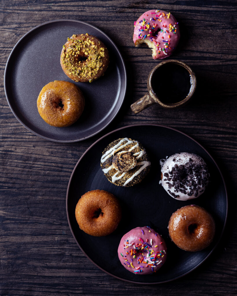 A variety of donuts sit on two black plates on a dark wood counter with a bitten donut and coffee cup sitting on the counter.