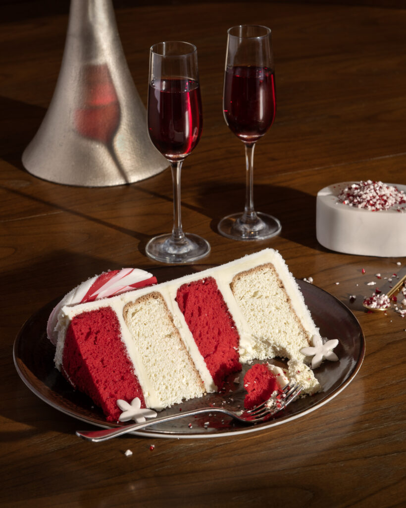 A slice of peppermint cake, white and red in color, sits on a plate next to two champagne glasses of red liquid. 