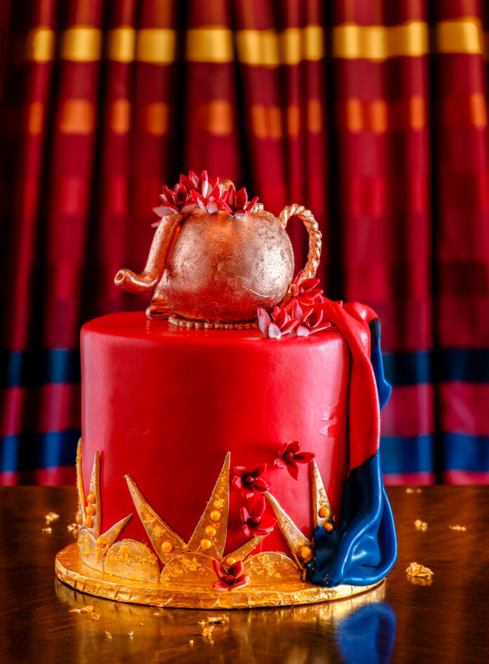 A colorful pink tiered cake with a sparkly pink tea pot on top with other yellow and blue decorations.