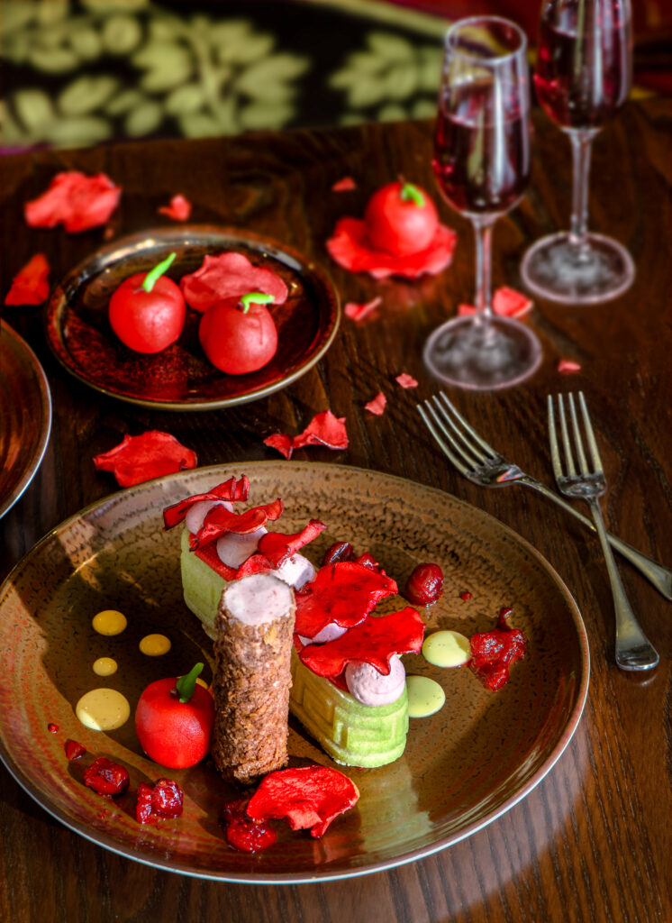 A brown plate with a very fancy red and green layered and decorated dessert, little marzipan apples on a smaller plate above and to the left, and champagne flutes on the right with a dark cranberry colored drink.