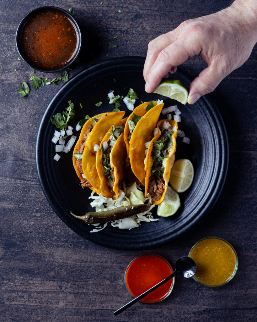 Birria tacos from a food truck in santa fe sit on a dark plate on a dark wood counter with a little bowl of Birria broth nearby and a spoon beneath the plate.