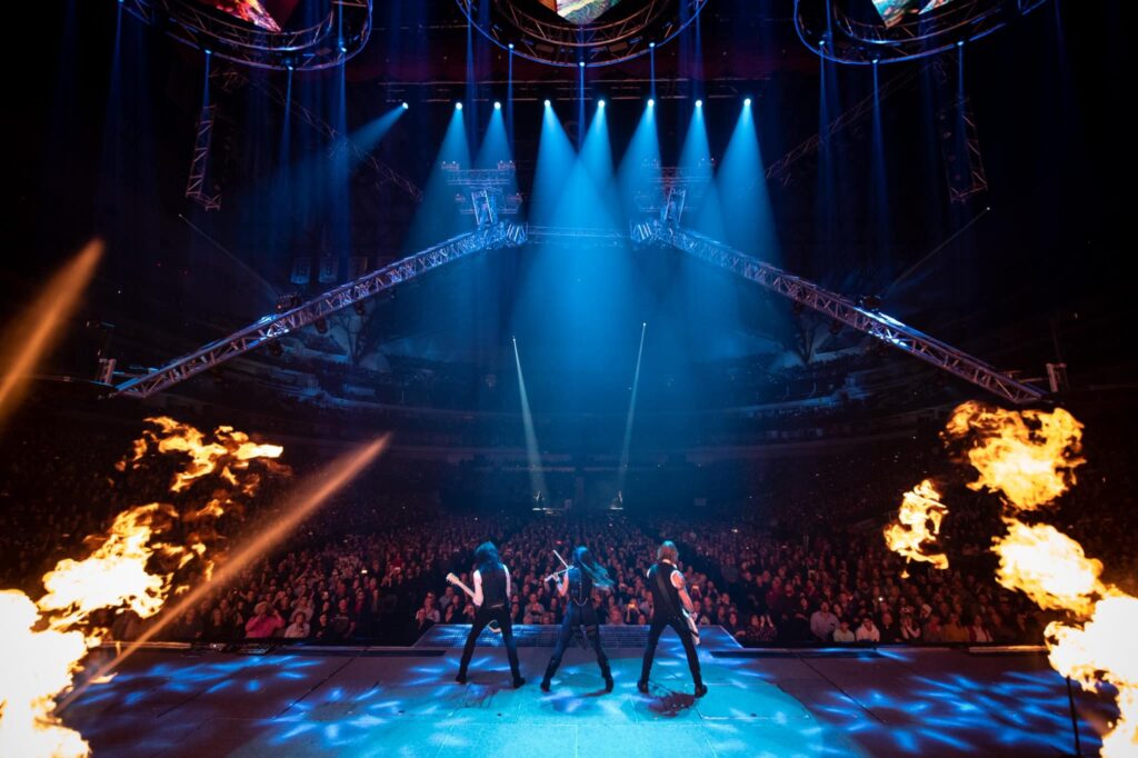 Three guitarists play on center stage under blue lights with a huge audience in front of them and fire on the sides as a promotion for Pittsburgh.