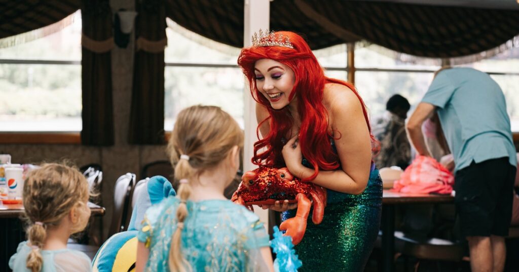 A woman dressed as Ariel from The LIttle Mermaid holds a crab stuffed animal and speaks to little girls in dresses.