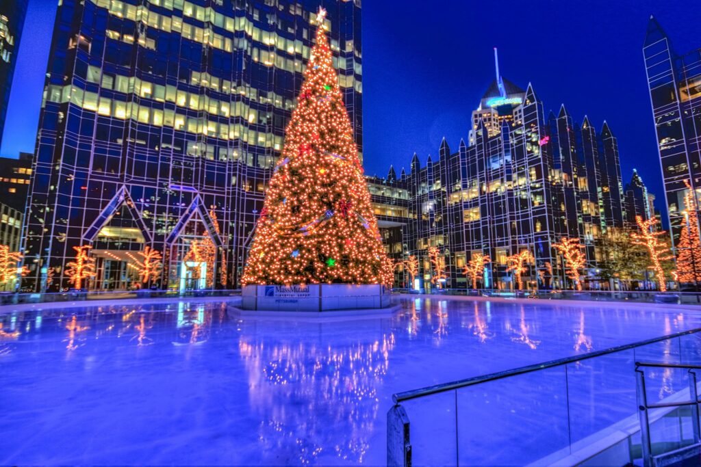 A Christmas tree lit up in the middle of an ice rink surrounded by city buildings and more lights.