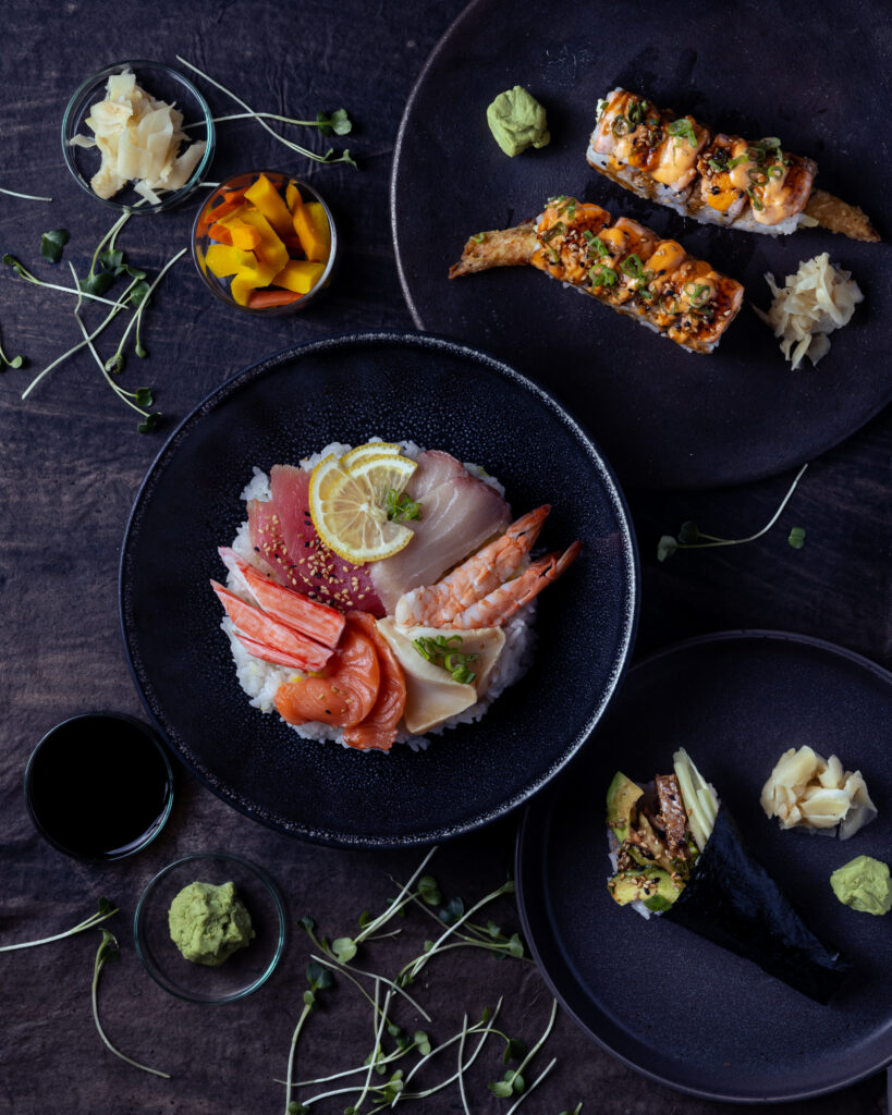 Raw fish, rice, and sushi from a food truck in Santa Fe is arranged on three different dark plates with sprigs of herbs in between the plates.