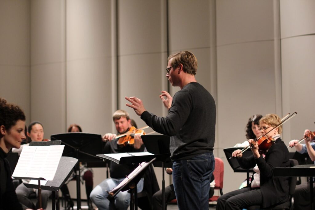 A conductor on a stand conducts different string players like violinists.