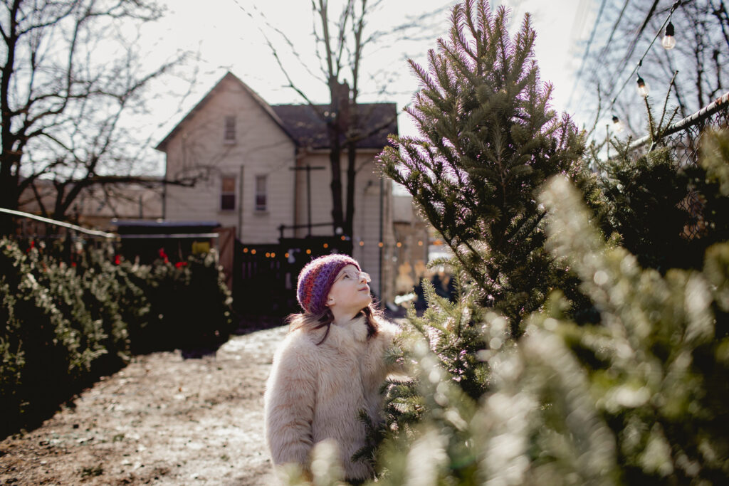 A girl in a beanie and glasses looks up at a tree at a tree farm.