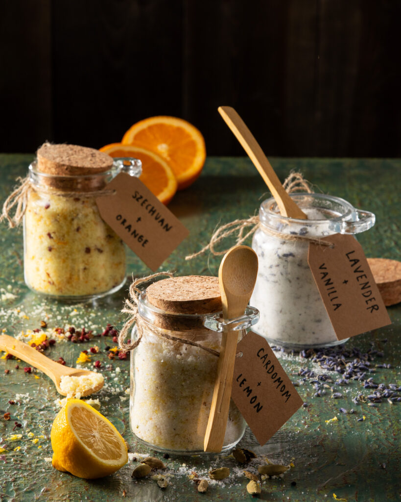Three homemade flavored sugars in small glass jars with cork lids , garnished with orange slices and cute little wooden spoons for serving,
