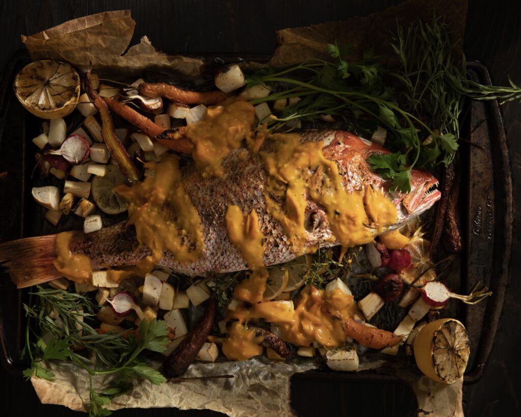 A roasted fish with a yellow sauce over roasted vegetables, garnished with fresh herbs.