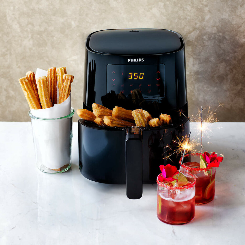 A black air fryer on a white counter with churros inside and cocktails beside the air fryer.