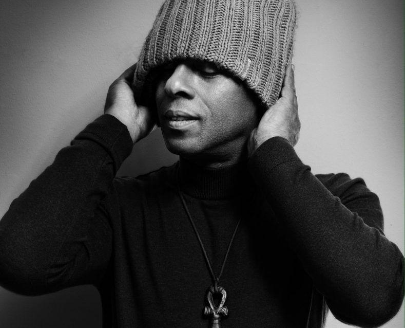 A black and white photo of a man in a beanie hat and a black turtleneck with a necklace on.