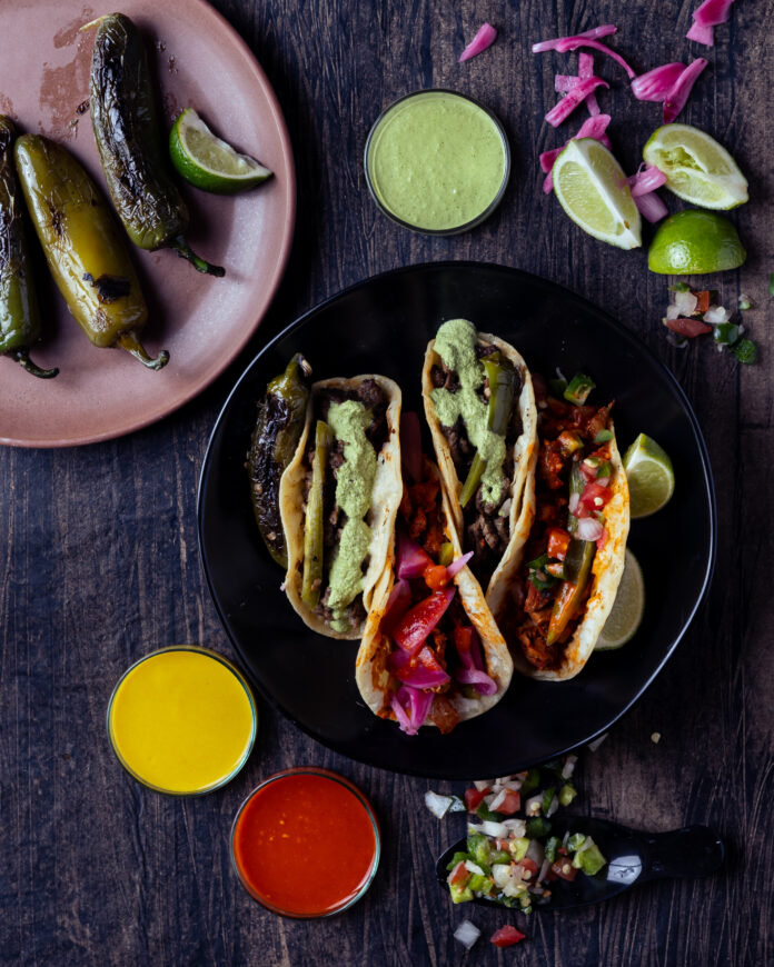 A black plate holds four tacos topped with green and purple sauce while a pink plate sits in the top left corner holding roasted peppers from a food truck in Santa Fe.