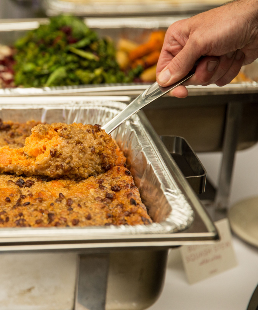 A hand slides in from the right of the frame and grabs a scoop of Bistro Pecan Sweet Potato Casserole from a foil serving tray.