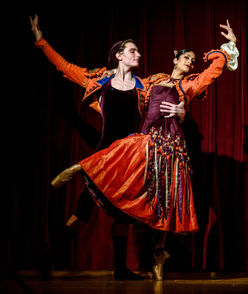 Two dancers, one man and one woman, hold each other with one arm and fling the other arm in the air. they are dressed in red and black outfits.