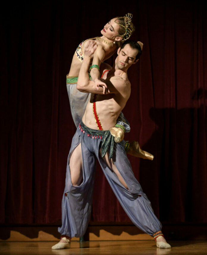 A male dancer lifts a female dancer into the air against his body on a stage.