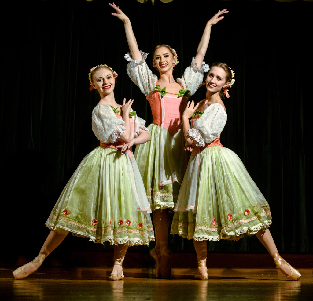 Three female ballet dancers wearing costume dresses of light green, pink and white.