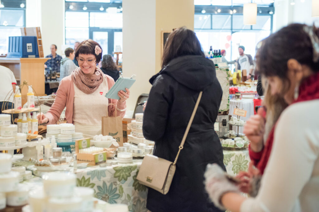 A woman stands behind a booth of homemade soaps with a smile on her face as she talks to another woman at the Pittsburgh market.