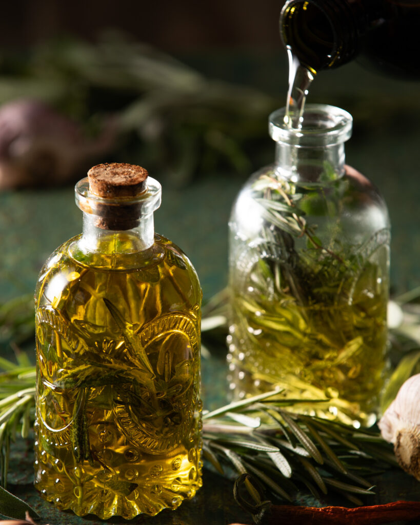 Two textured clear glass bottles with cork closure, filled with homemade herb-infused olive oil made with dried rosemary, thyme, and oregano.