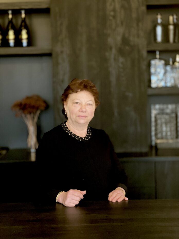 An older woman with short brownish-red hair sits at a brown kitchen table.