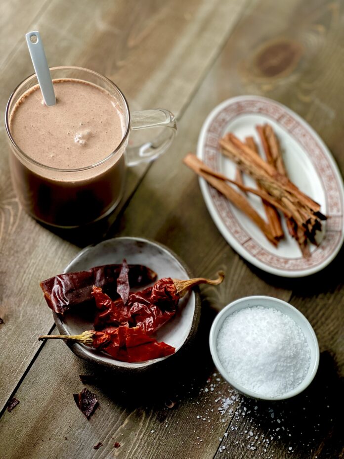 A clear glass mug of spiked hot chocolate with a small plate of dried red ancho chilis, a small oval dish of cinnamon sticks, and a small bowl of salt on a wooden surface