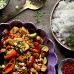 A purple scalloped serving dish with a chicken and brightly colored vegetable mix with a bowl of rice in the upper right corner on a brown wooden surface.