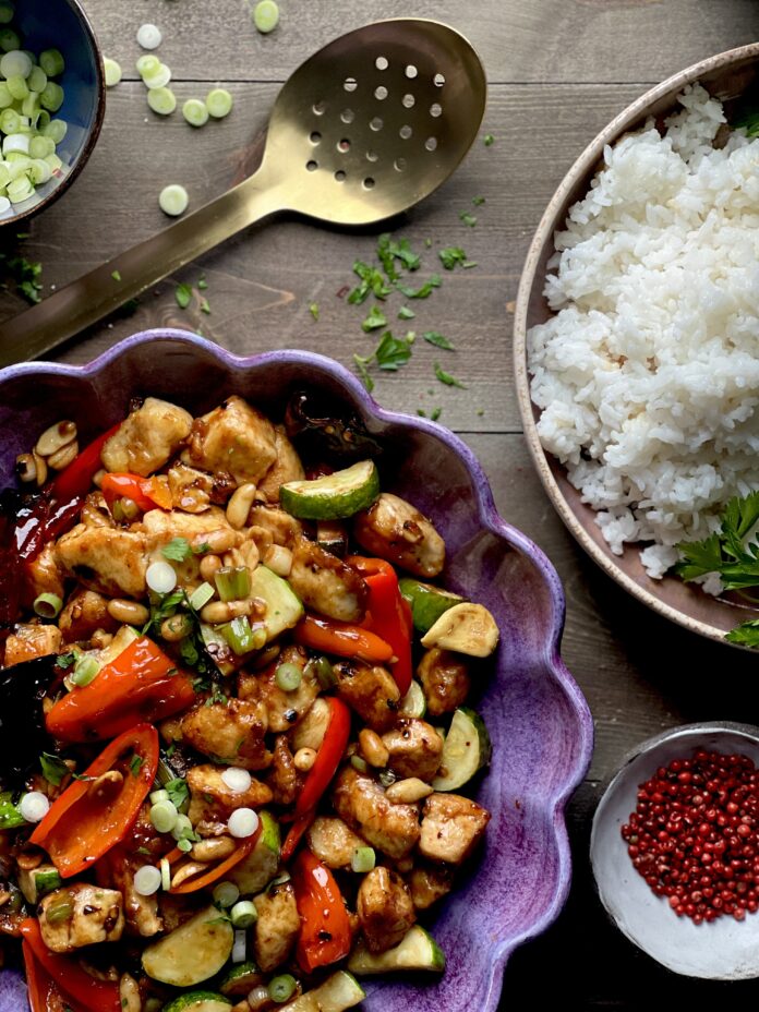 A purple scalloped serving dish with a chicken and brightly colored vegetable mix with a bowl of rice in the upper right corner on a brown wooden surface.