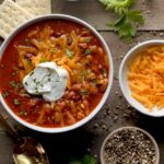 An overhead view of The best Wendy's Copycat chili in a bowl with grated cheddar cheese and a dollop of sour cream in a white bowl, with small bowls of sour cream, grated cheddar, and pepper, a couple saltine crackers, parsley sprigs and a gold spoon on a wooden surface.