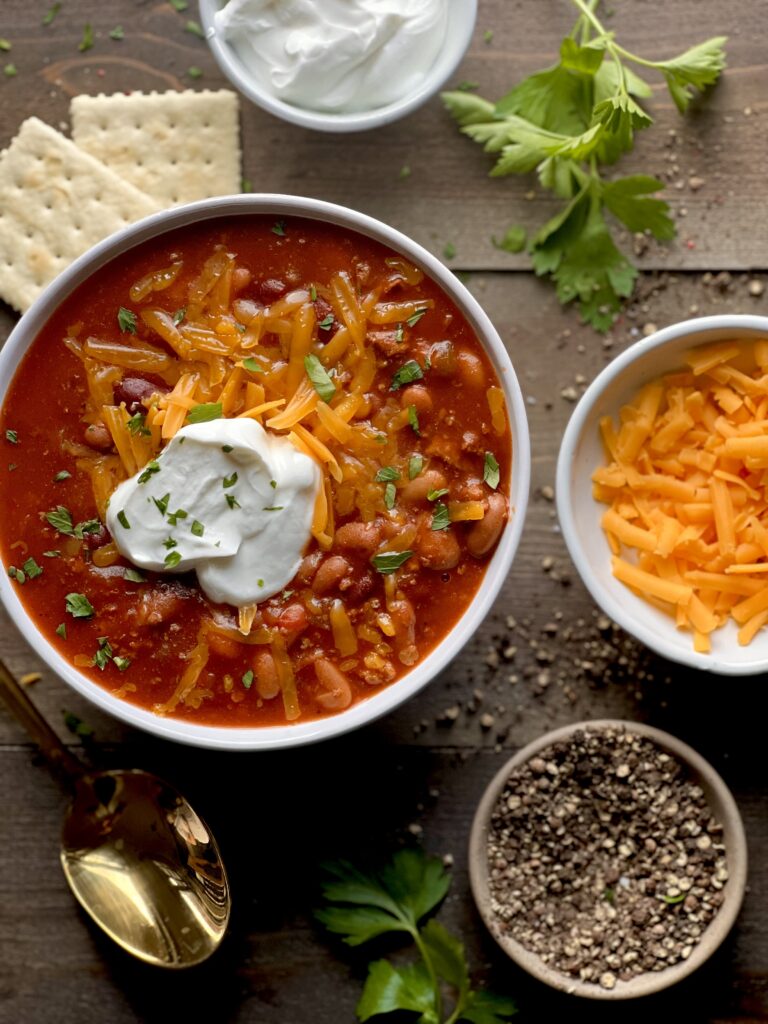 An overhead view of The best Wendy's Copycat chili in a bowl with grated cheddar cheese and a dollop of sour cream in a white bowl, with small bowls of sour cream, grated cheddar, and pepper, a couple saltine crackers, parsley sprigs and a gold spoon on a wooden surface.