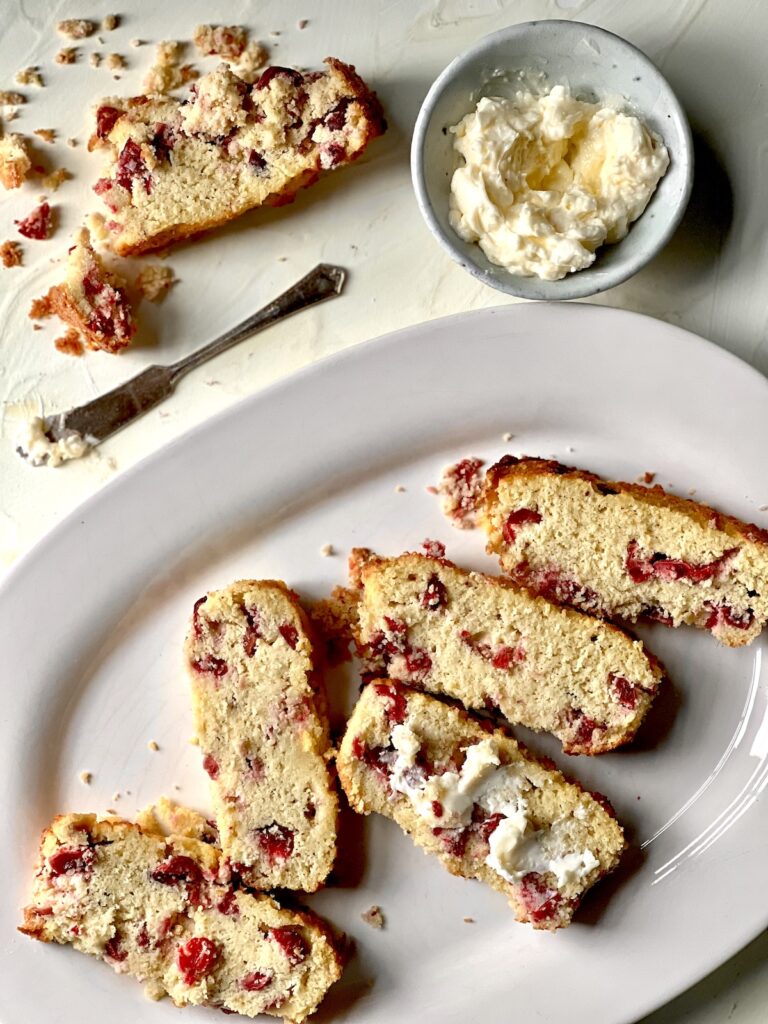 An aerial view of a lemon keto cranberry quick bread sitting chopped up into five slices on a white plate. the middle slice has butter on it. A small bowl of whipped butter sits to the top right.
