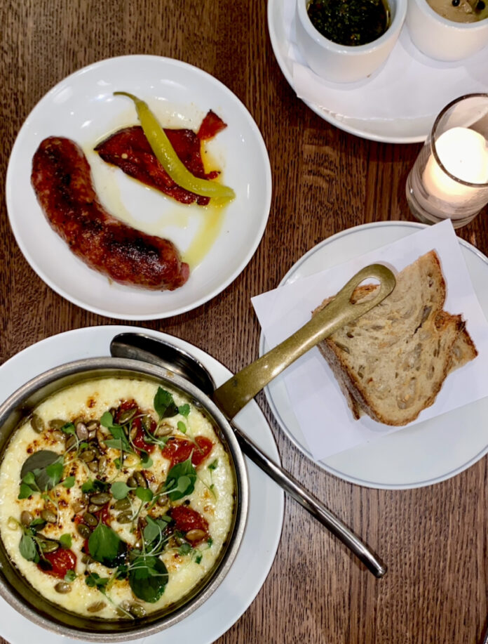 A cheese skillet, sausage, and bread sit on a dining table with chimichurri in reach at Balvanera Pittsburgh.