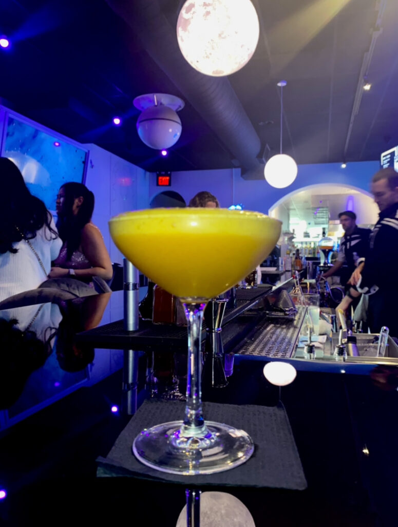 An orange and yellow cocktail sits in a glass on a reflective black bar with moon light orbs above at Space Bar in Market Square.