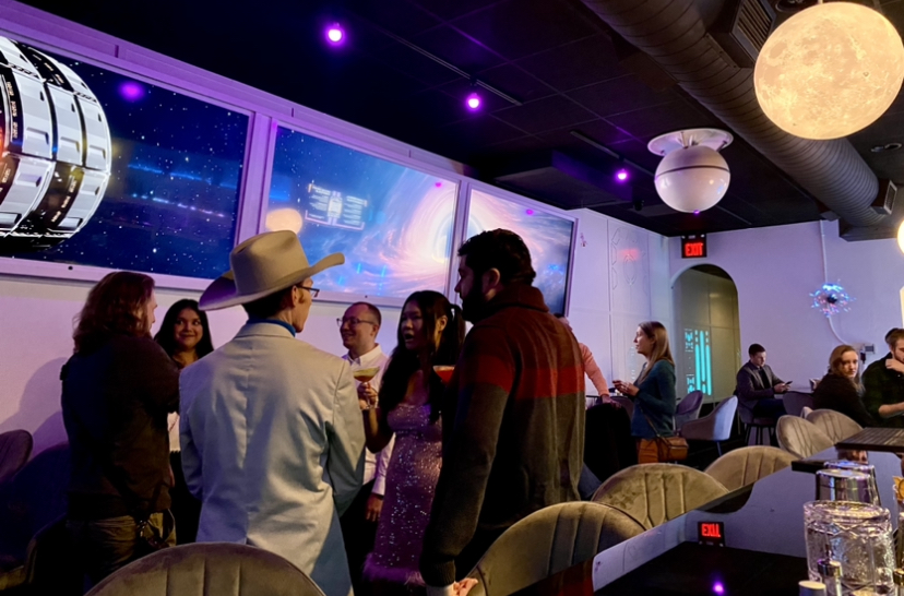 A space bar decorated in tv screens displaying images of the galaxy with people talking throughout the space in Space Bar at Market Square.